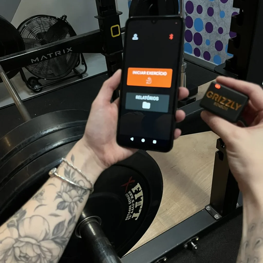 Grizzly Sensor ao lado de um smartphone com o aplicativo Grizzly aberto, em uma academia